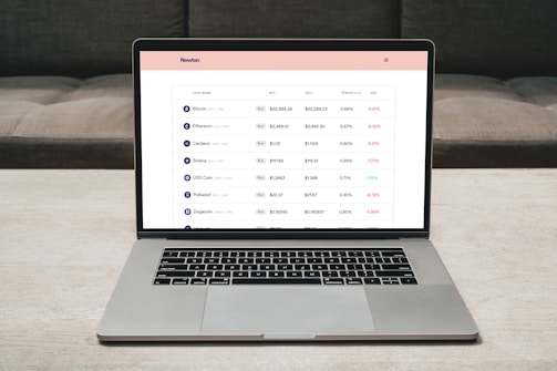 A laptop on a table displaying a cryptocurrency trading platform. The screen lists various cryptocurrencies along with their buy and sell values, spread percentages, and 24-hour change percentages. The keyboard and touchpad of the laptop are visible, set against a neutral background with a sofa in the back.