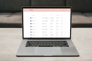 A laptop on a table displaying a cryptocurrency trading platform. The screen lists various cryptocurrencies along with their buy and sell values, spread percentages, and 24-hour change percentages. The keyboard and touchpad of the laptop are visible, set against a neutral background with a sofa in the back.