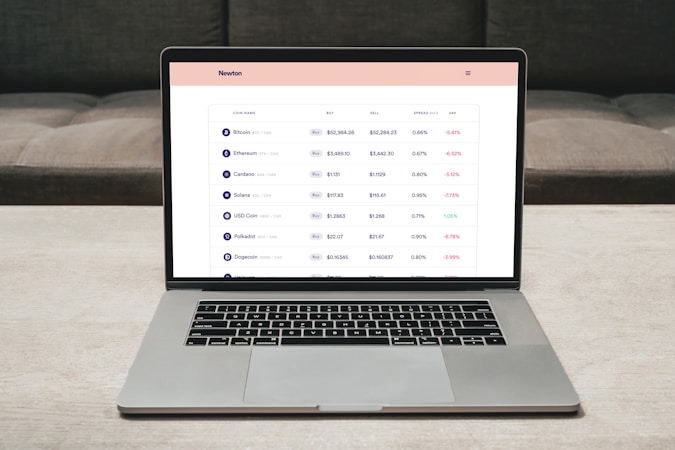 A laptop on a table displaying a cryptocurrency trading platform. The screen lists various cryptocurrencies along with their buy and sell values, spread percentages, and 24-hour change percentages. The keyboard and touchpad of the laptop are visible, set against a neutral background with a sofa in the back.