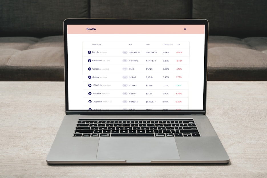 A laptop on a table displaying a cryptocurrency trading platform. The screen lists various cryptocurrencies along with their buy and sell values, spread percentages, and 24-hour change percentages. The keyboard and touchpad of the laptop are visible, set against a neutral background with a sofa in the back.