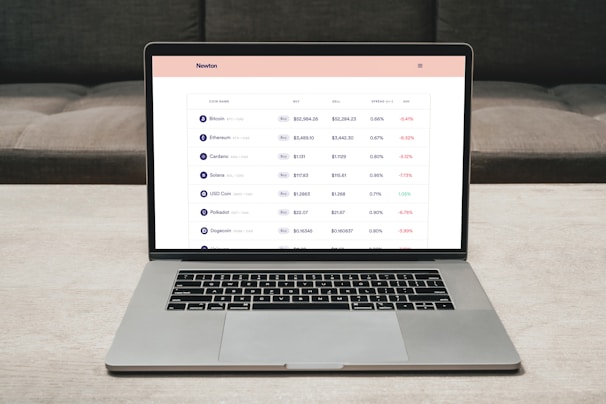 A laptop on a table displaying a cryptocurrency trading platform. The screen lists various cryptocurrencies along with their buy and sell values, spread percentages, and 24-hour change percentages. The keyboard and touchpad of the laptop are visible, set against a neutral background with a sofa in the back.