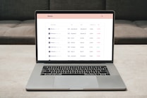 A laptop on a table displaying a cryptocurrency trading platform. The screen lists various cryptocurrencies along with their buy and sell values, spread percentages, and 24-hour change percentages. The keyboard and touchpad of the laptop are visible, set against a neutral background with a sofa in the back.