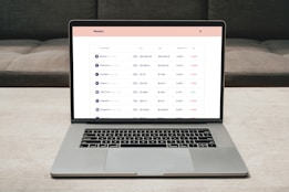 A laptop on a table displaying a cryptocurrency trading platform. The screen lists various cryptocurrencies along with their buy and sell values, spread percentages, and 24-hour change percentages. The keyboard and touchpad of the laptop are visible, set against a neutral background with a sofa in the back.