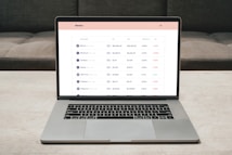 A laptop on a table displaying a cryptocurrency trading platform. The screen lists various cryptocurrencies along with their buy and sell values, spread percentages, and 24-hour change percentages. The keyboard and touchpad of the laptop are visible, set against a neutral background with a sofa in the back.