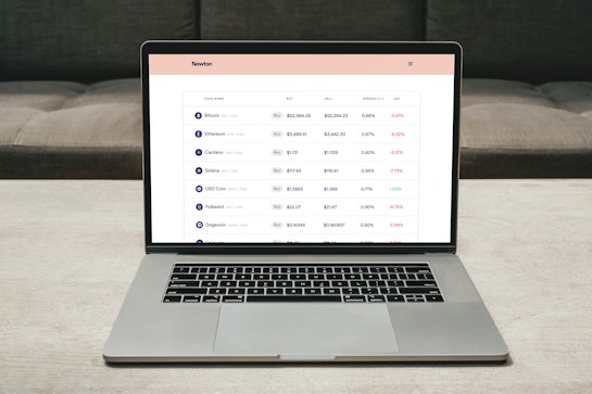 A laptop on a table displaying a cryptocurrency trading platform. The screen lists various cryptocurrencies along with their buy and sell values, spread percentages, and 24-hour change percentages. The keyboard and touchpad of the laptop are visible, set against a neutral background with a sofa in the back.