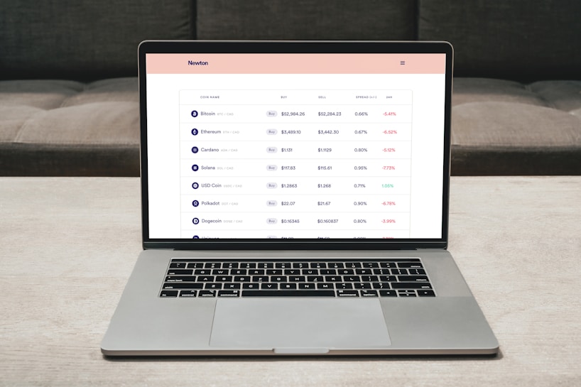 A laptop on a table displaying a cryptocurrency trading platform. The screen lists various cryptocurrencies along with their buy and sell values, spread percentages, and 24-hour change percentages. The keyboard and touchpad of the laptop are visible, set against a neutral background with a sofa in the back.