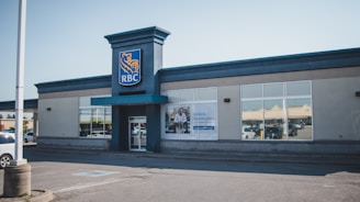 A single-story building with a prominent blue and yellow logo is visible. The entrance features large glass windows and a blue awning. A poster is displayed on one of the windows, and the surrounding environment includes a parking lot with a few parked cars.
