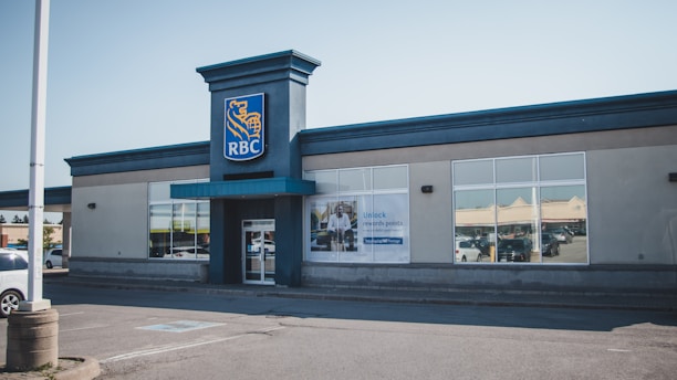 A single-story building with a prominent blue and yellow logo is visible. The entrance features large glass windows and a blue awning. A poster is displayed on one of the windows, and the surrounding environment includes a parking lot with a few parked cars.