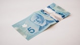 A Canadian five-dollar bill lies on a plain surface, displaying the portrait of a notable figure in shades of blue. The bill features elaborate designs, including a large numeral '5', maple leaf motifs, and transparent elements. The background is neutral, emphasizing the vibrant colors and intricate details of the currency.