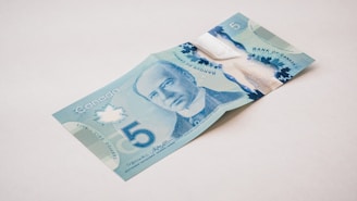 A Canadian five-dollar bill lies on a plain surface, displaying the portrait of a notable figure in shades of blue. The bill features elaborate designs, including a large numeral '5', maple leaf motifs, and transparent elements. The background is neutral, emphasizing the vibrant colors and intricate details of the currency.
