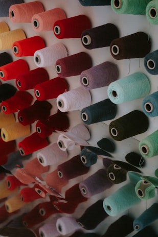 Spools of thread in various vibrant colors are neatly arranged in rows on a display wall. There are multiple colors including red, pink, yellow, teal, and others. The threads have a soft texture and are reflected on a glossy surface below them.