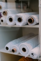 Several large rolls of white fabric are neatly arranged on white shelves. The rolls have visible cardboard tubes at their centers and vary slightly in texture. They appear organized and ready for use, suggesting a warehouse or fabric store setting.