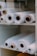 Several large rolls of white fabric are neatly arranged on white shelves. The rolls have visible cardboard tubes at their centers and vary slightly in texture. They appear organized and ready for use, suggesting a warehouse or fabric store setting.
