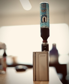 A vape device sits on a table with a blue slushy flavored e-liquid bottle attached to the top. The branding on the device reads 'SMOK' and the drip tip is labeled 'DROP'. The background is blurred, giving focus to the vape and bottle.