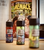 Three bottles of e-liquid are placed on a table with colorful labels, in front of a comic book featuring bold yellow text and illustrative artwork. A blurred background includes pages of a book and a computer screen.