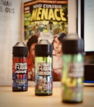 Three bottles of e-liquid are placed on a table with colorful labels, in front of a comic book featuring bold yellow text and illustrative artwork. A blurred background includes pages of a book and a computer screen.