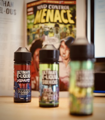Three bottles of e-liquid are placed on a table with colorful labels, in front of a comic book featuring bold yellow text and illustrative artwork. A blurred background includes pages of a book and a computer screen.