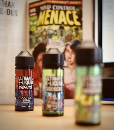 Three bottles of e-liquid are placed on a table with colorful labels, in front of a comic book featuring bold yellow text and illustrative artwork. A blurred background includes pages of a book and a computer screen.