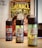 Three bottles of e-liquid are placed on a table with colorful labels, in front of a comic book featuring bold yellow text and illustrative artwork. A blurred background includes pages of a book and a computer screen.