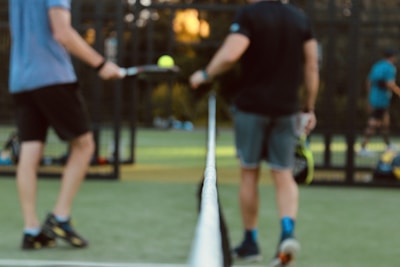 Two individuals holding tennis rackets are standing on either side of a tennis net on a court. The focus is on the net, with the players blurred in the background. One person is wearing blue and black sports attire, while the other wears black and grey. Tennis balls and additional players can be seen further in the background.