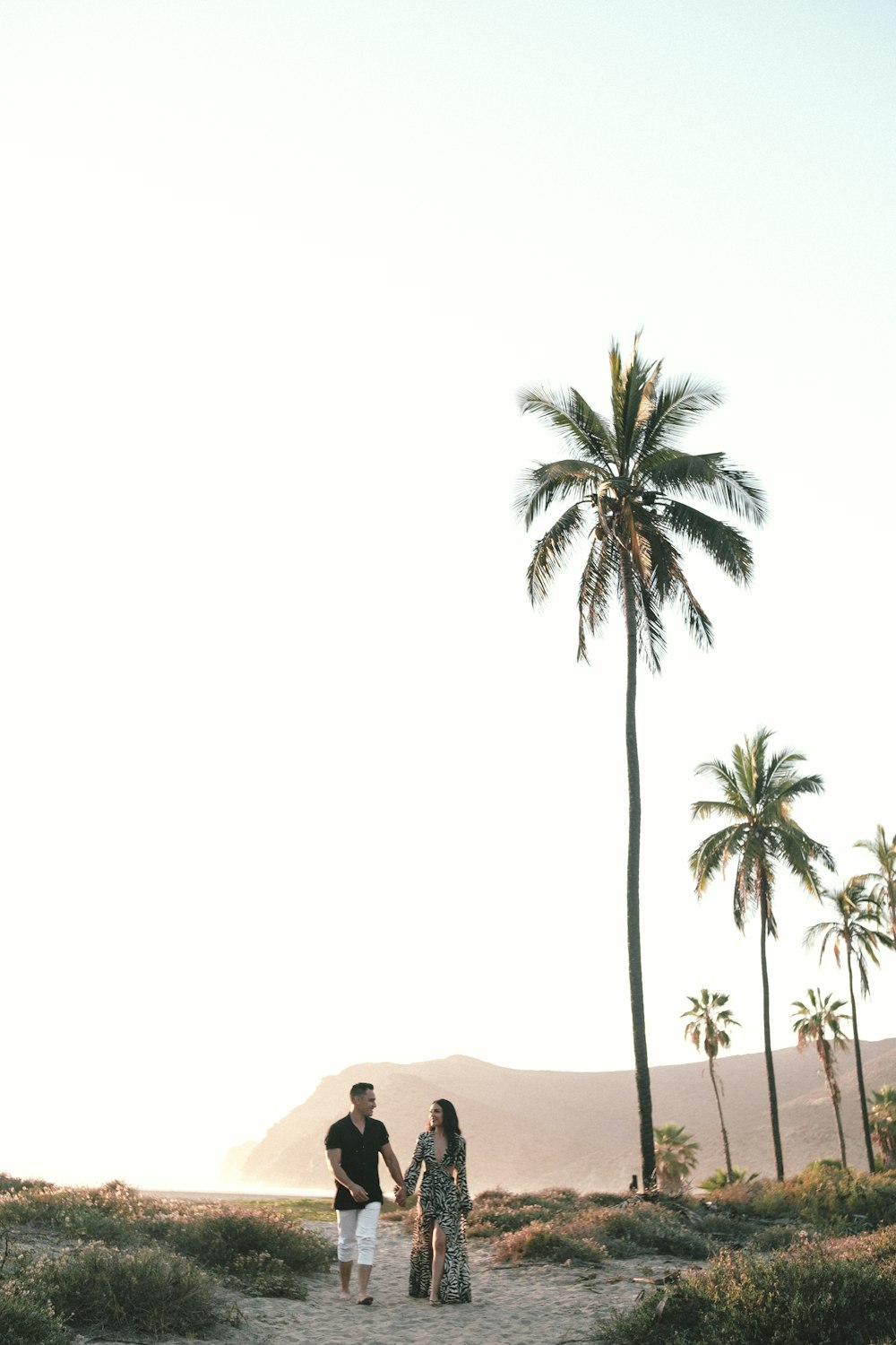 a man and woman walking on a path with palm trees and sand and mountains in the background