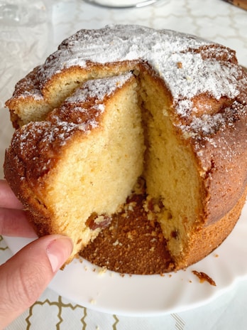 A freshly baked round cake with a slice cut out, revealing a soft, fluffy interior. The cake is dusted with powdered sugar, adding a decorative and sweet touch. It sits on a white plate, and a hand is holding one side of the cake, ready to serve or take a piece.