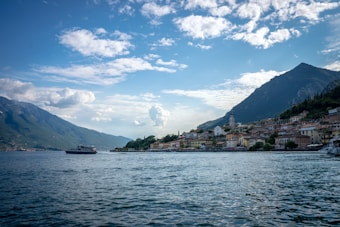 A picturesque lakeside town with a serene body of water in the foreground, surrounded by mountains. The town features charming architecture with a prominent church steeple. The sky is partly cloudy with a bright, crisp atmosphere. A boat can be seen moving across the water, contributing to the tranquil scenery.