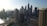 A panoramic view of a bustling cityscape with numerous tall skyscrapers. The iconic Burj Khalifa stands prominently against a clear blue sky. A curving waterfront and bridge are visible, and construction cranes can be seen atop some of the buildings, indicating ongoing development.