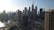 A panoramic view of a bustling cityscape with numerous tall skyscrapers. The iconic Burj Khalifa stands prominently against a clear blue sky. A curving waterfront and bridge are visible, and construction cranes can be seen atop some of the buildings, indicating ongoing development.