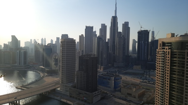 A panoramic view of a bustling cityscape with numerous tall skyscrapers. The iconic Burj Khalifa stands prominently against a clear blue sky. A curving waterfront and bridge are visible, and construction cranes can be seen atop some of the buildings, indicating ongoing development.