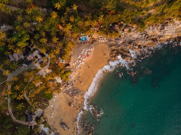 An aerial view of a scenic beach surrounded by lush palm trees and rocky cliffs. The sandy shore is equipped with numerous sun loungers and beach umbrellas, some of which are red. The turquoise ocean waves gently lap against the shoreline. Small structures and pathways are visible amidst the dense greenery along the coast.