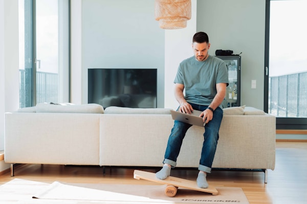 A man sitting on the backrest of a sofa while using a laptop. He balances on a wooden balance board, creating a sense of focus and multitasking. The living room is modern and bright, featuring a large window, a television, and minimalistic decor.