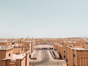 A wide, sunlit view of a desert cityscape with light-colored, flat-roofed buildings lined up in structured rows. The sky is clear and slightly tinted, indicating a warm climate. Satellite dishes are visible on the rooftops, and empty streets run between the buildings, extending toward the horizon with a hazy skyline in the distance.