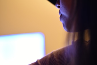 A person is partially visible, facing a bright screen that casts a glow. The focus is on their side profile near a glowing monitor in a dimly lit setting.