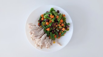A white plate contains a serving of shredded chicken and a colorful mix of vegetables, including sliced green beans, diced carrots, kidney beans, and corn kernels, arranged in a neat presentation.