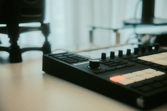 A music production device with a series of buttons and knobs sits on a table, surrounded by a softly blurred background. The foreground is in focus highlighting the sleek design of the equipment. A bright light from a nearby window softly illuminates the scene, casting a gentle glow over the dark surface of the device.
