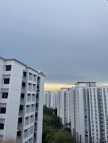 A cluster of high-rise residential buildings with white facades and pink accents under an overcast sky. There is a patch of greenery visible in the lower part of the image.