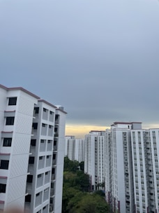 A cluster of high-rise residential buildings with white facades and pink accents under an overcast sky. There is a patch of greenery visible in the lower part of the image.