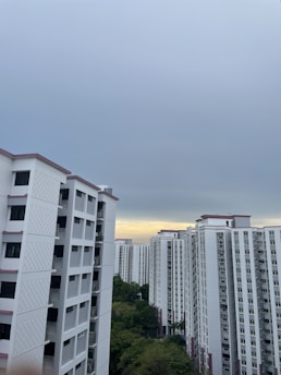A cluster of high-rise residential buildings with white facades and pink accents under an overcast sky. There is a patch of greenery visible in the lower part of the image.