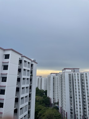 A cluster of high-rise residential buildings with white facades and pink accents under an overcast sky. There is a patch of greenery visible in the lower part of the image.