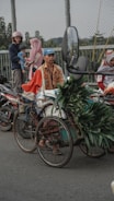 A man wearing a blue cap and a patterned shirt rides a bicycle that has been modified to carry cargo. The cargo includes bundles of green leafy plants. The bicycle is parked on a street beside a fence. In the background, a family with children, wearing helmets and headscarves, is seen standing near two motorcycles.