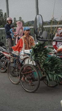 A man wearing a blue cap and a patterned shirt rides a bicycle that has been modified to carry cargo. The cargo includes bundles of green leafy plants. The bicycle is parked on a street beside a fence. In the background, a family with children, wearing helmets and headscarves, is seen standing near two motorcycles.