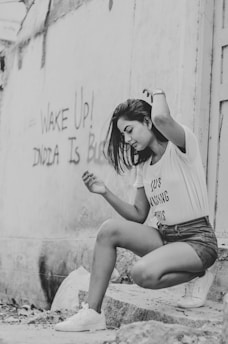 A young woman with long hair is captured in a candid pose against an urban backdrop. She is wearing a casual outfit consisting of a T-shirt and shorts. Graffiti on the wall in the background reads 'Wake Up! India Is'. The image is in black and white, adding a dramatic and timeless feel.