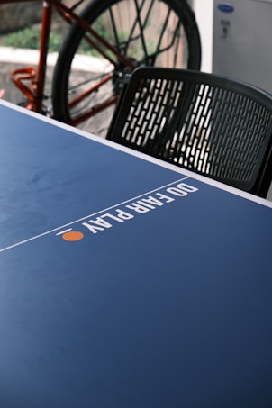 A close-up view of a blue ping pong table with the words 'DO EPIC PLAY' printed on it. In the background, part of a bicycle is visible with a focus on the wheel. A black plastic chair is positioned nearby.