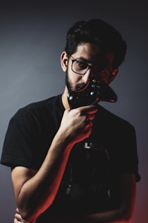 A person with glasses holds a video game controller close to their face against a dark background. The lighting casts dramatic shadows, emphasizing features and creating a moody atmosphere.