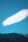 A large, white cloud with a smooth, elongated shape floats above a range of dark green mountains under a bright blue sky.