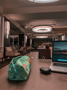 A modern indoor space with a sophisticated look featuring seating areas with beige armchairs and side tables. Large circular ceiling lights illuminate the area, and a wooden desk in the foreground holds a colorful pencil case, a black wireless mouse, and an open laptop displaying a scenic wallpaper.