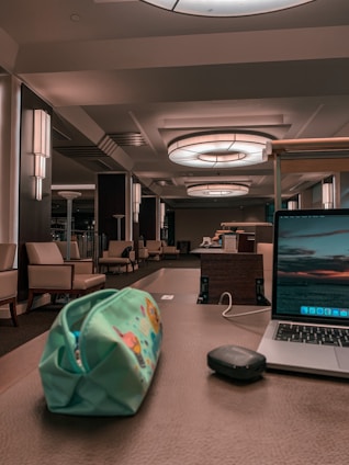 A modern indoor space with a sophisticated look featuring seating areas with beige armchairs and side tables. Large circular ceiling lights illuminate the area, and a wooden desk in the foreground holds a colorful pencil case, a black wireless mouse, and an open laptop displaying a scenic wallpaper.