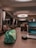 A modern indoor space with a sophisticated look featuring seating areas with beige armchairs and side tables. Large circular ceiling lights illuminate the area, and a wooden desk in the foreground holds a colorful pencil case, a black wireless mouse, and an open laptop displaying a scenic wallpaper.