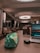 A modern indoor space with a sophisticated look featuring seating areas with beige armchairs and side tables. Large circular ceiling lights illuminate the area, and a wooden desk in the foreground holds a colorful pencil case, a black wireless mouse, and an open laptop displaying a scenic wallpaper.
